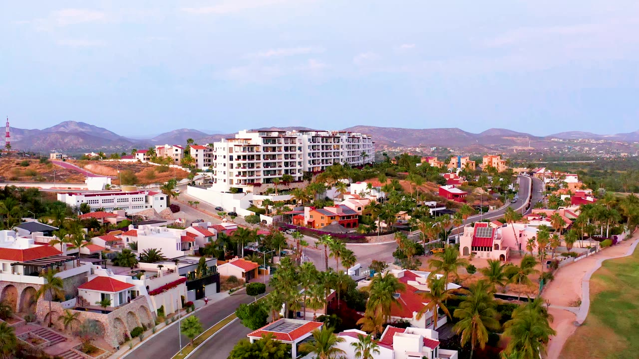 Second frame over San Jose del Cabo BCS Mexico, showing the road curving through palms and low buildings while a large white apartment block sits below the mountain horizon, with no visible people.
