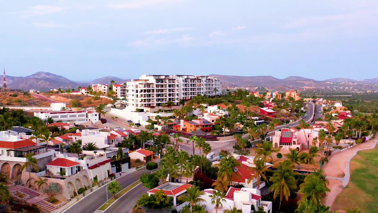 Third frame of the San Jose del Cabo residential area, with palms filling the foreground, red-roofed homes and white walls in the midground, and dry hills behind, with no visible people.