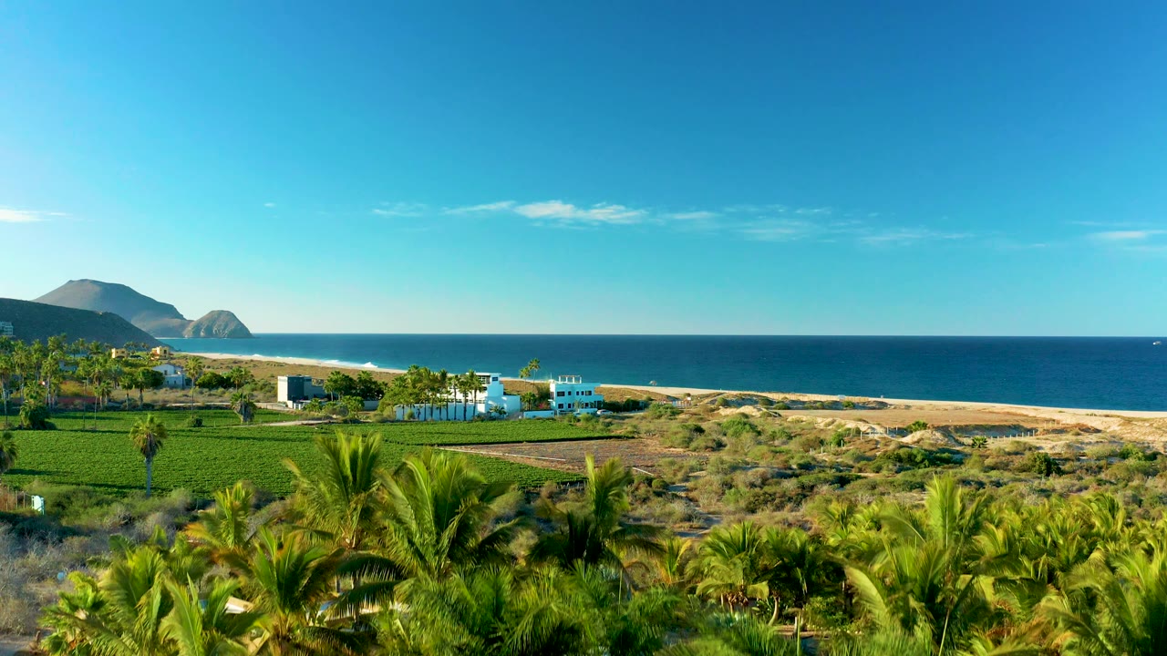 Beachfront fields, white buildings, ocean, and distant hills under clear sky.
