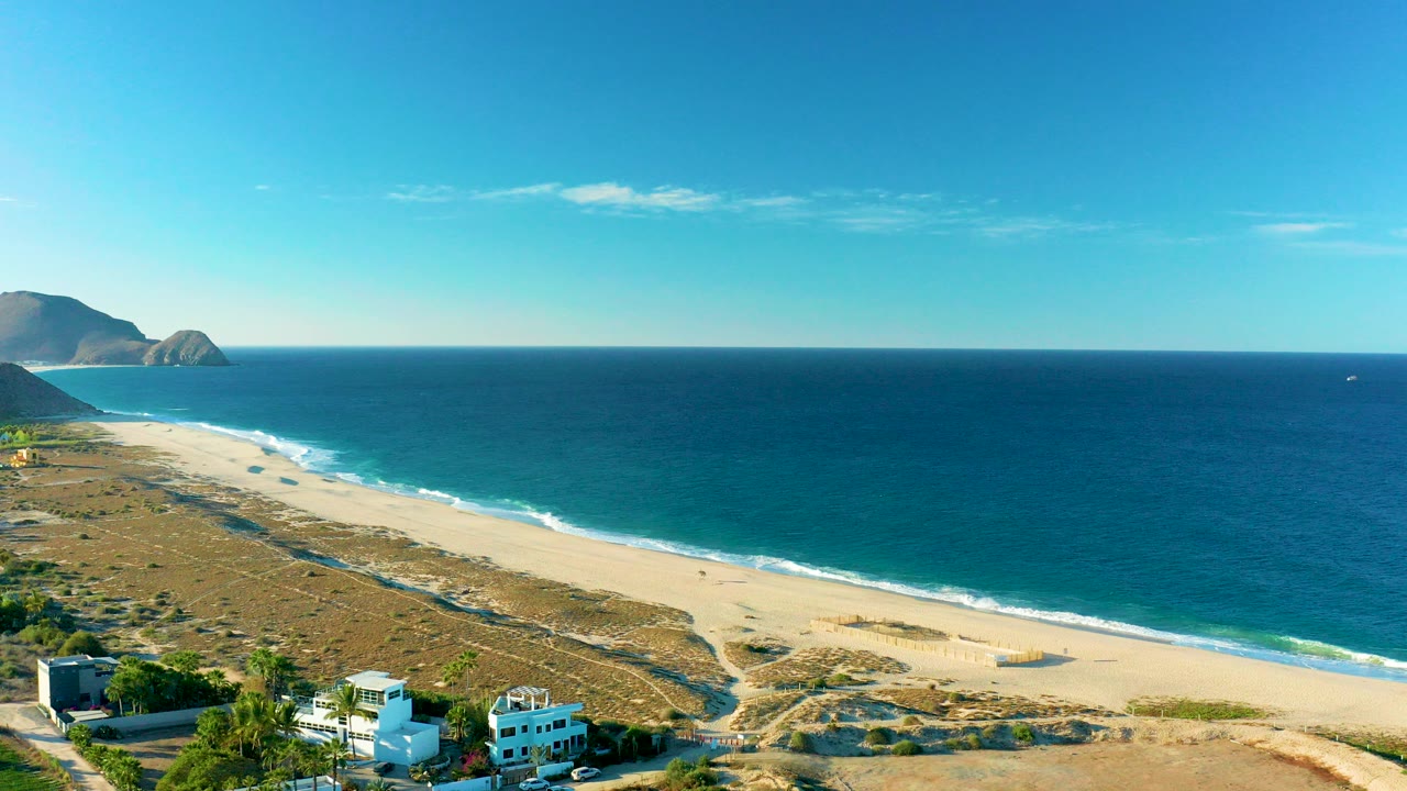 Opening aerial frame over Todos Santos, Baja California Sur, with foreground buildings below, sandy dunes, a long beach, blue ocean, distant hills, and no visible people.