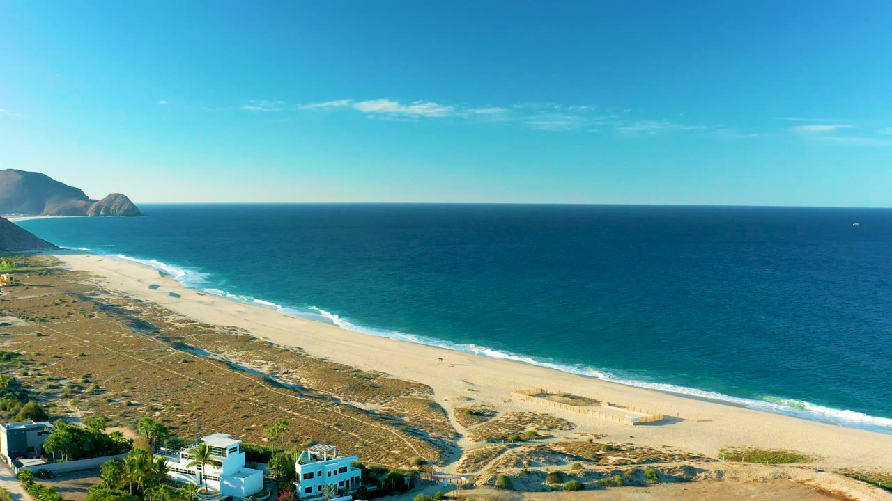 Todos Santos aerial coastline frame with the beach running diagonally below blue water, low dunes inland, distant hills at left, a small boat offshore, and no visible people.