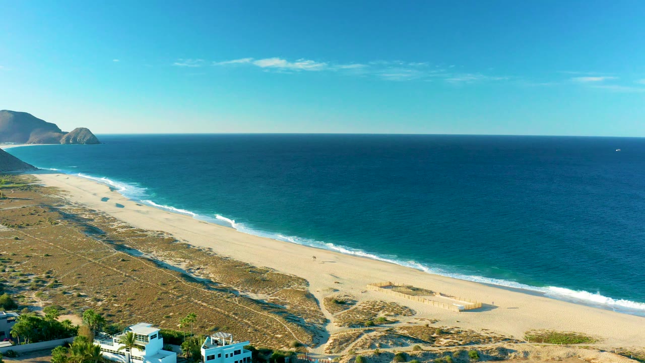 Beach and ocean aerial view with dunes, surf lines, distant hills, and buildings partly visible at the bottom.