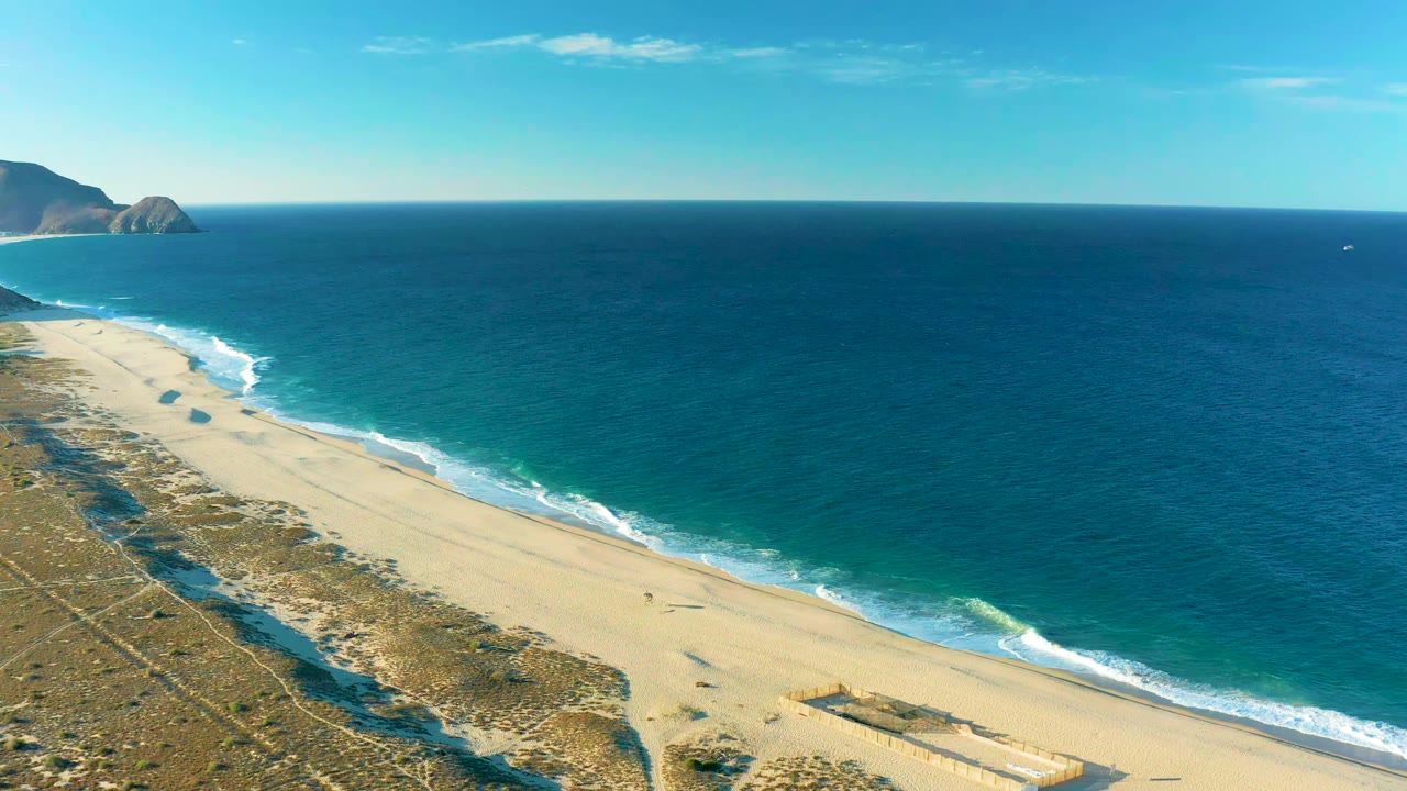 Aerial view over Todos Santos shoreline with the beach running diagonally across frame, white foam at the waterline, blue ocean beyond, and no visible people.