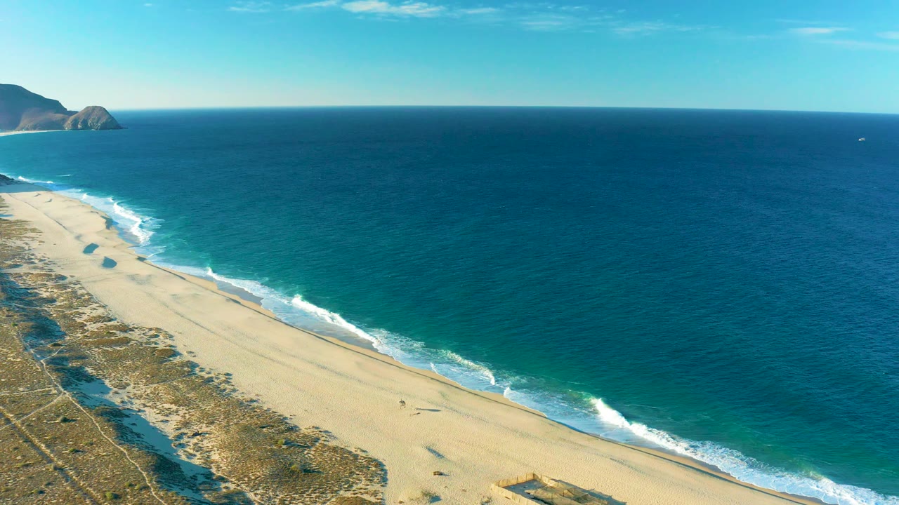 Aerial frame from Todos Santos showing more open ocean across the right side, beach and dunes along the left edge, distant rocks, and no visible people.