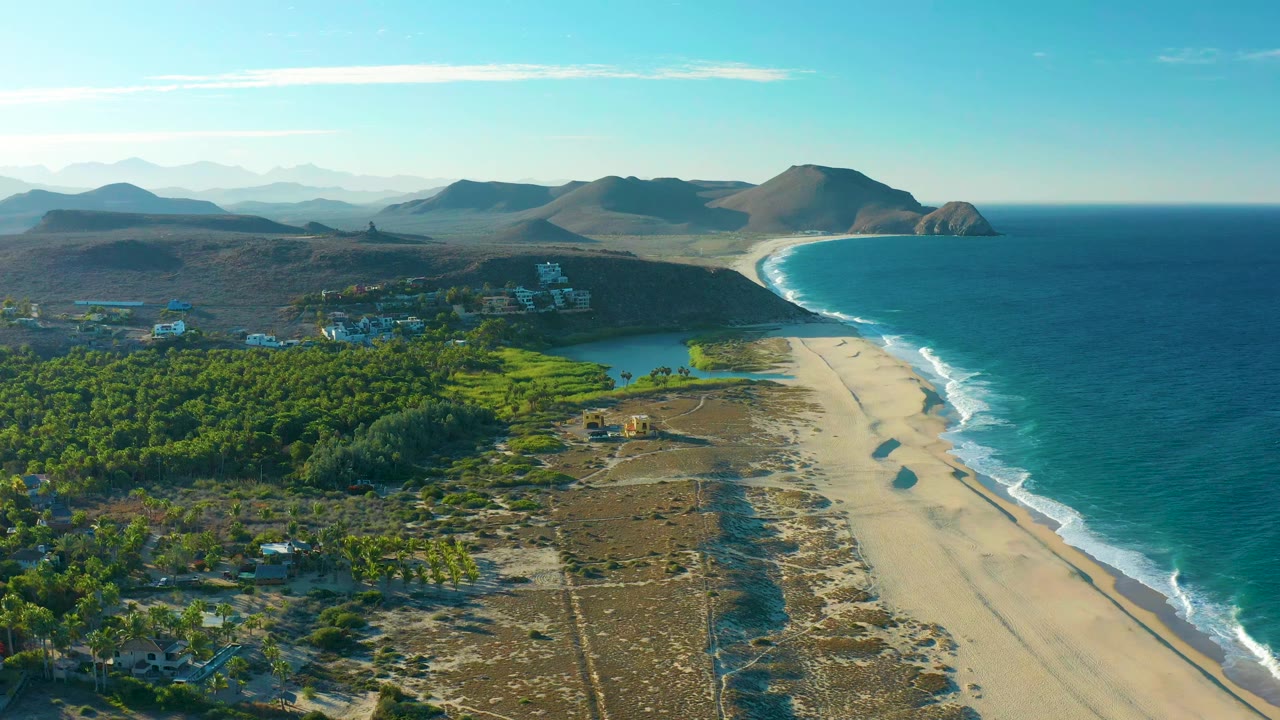 Aerial view one second into the Todos Santos clip, showing the lagoon just inland from the sandy beach, blue ocean and surf to the right, green vegetation and small structures below, distant hills, and no visible people.