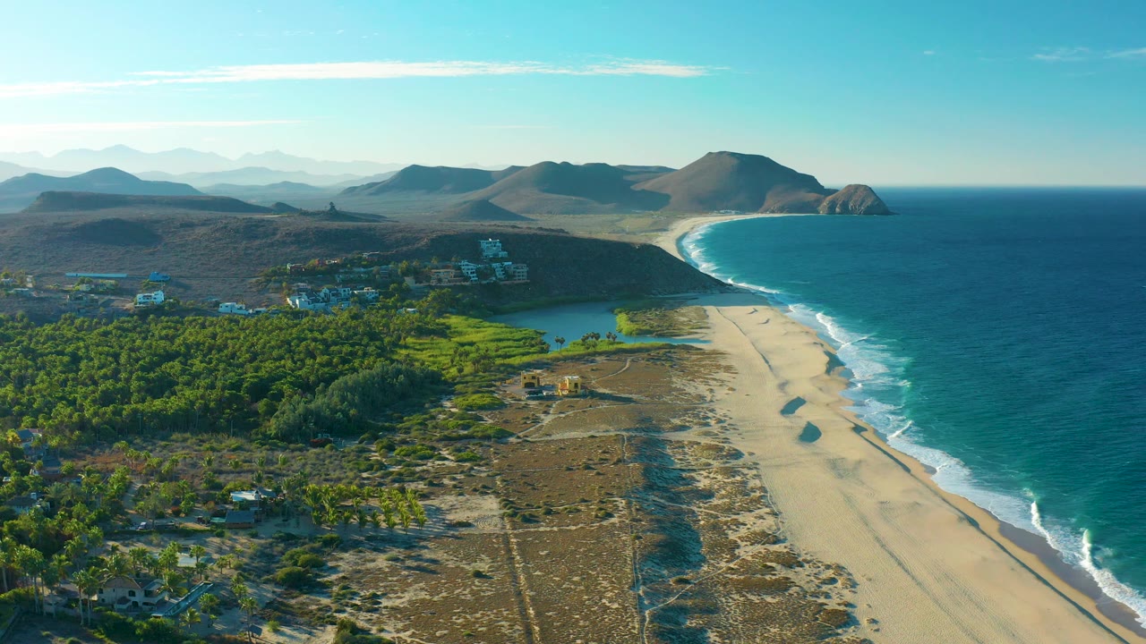 Aerial coastline with green vegetation, lagoon, sandy beach, waves, and hills.