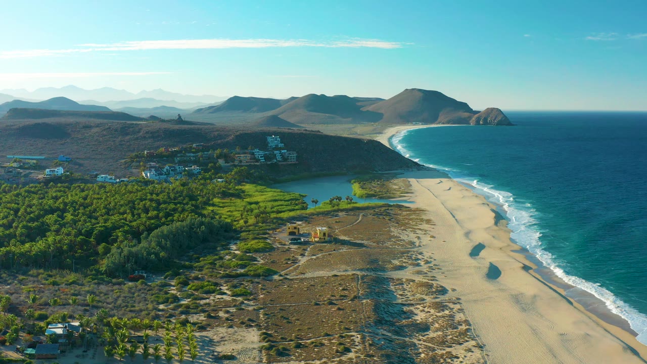 Aerial view at 0:01 of the Todos Santos shoreline with white surf tracing the beach, dense greenery inland, low hills in the distance, and no visible people.