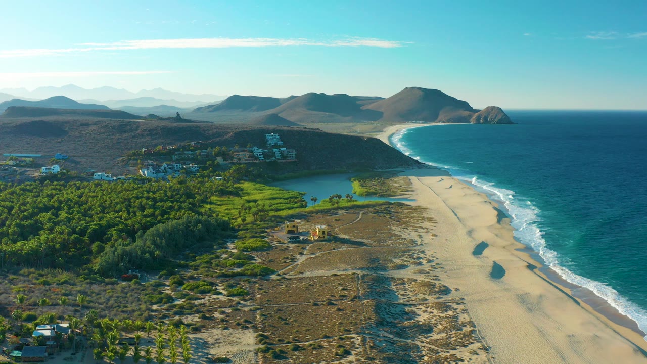 Aerial frame at 0:02 showing the Todos Santos beach curving past dunes and a small lagoon toward distant hills, with no visible people.