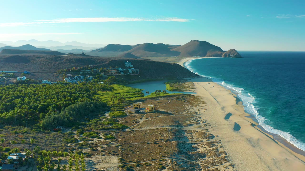 Mid-clip aerial view of the Todos Santos coast with sandy shore, rolling surf, green inland vegetation, scattered buildings, distant hills, and no visible people.