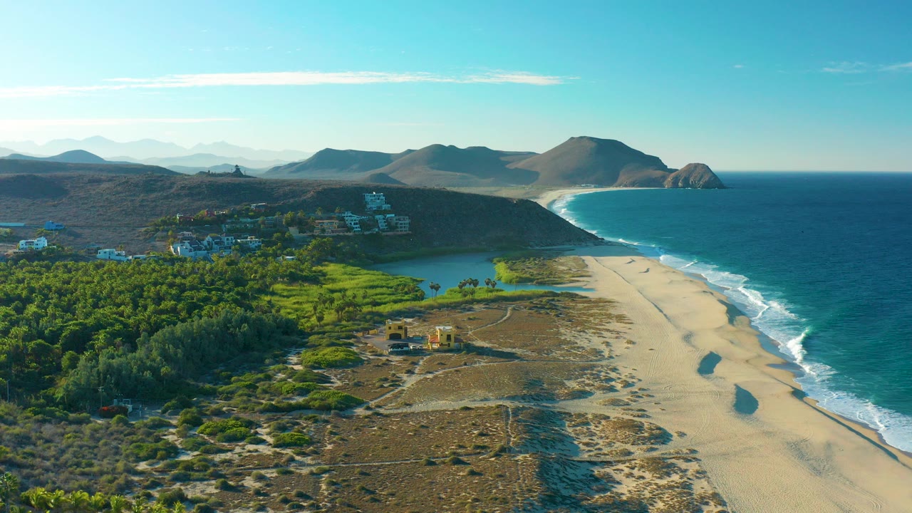 Opening aerial view over Todos Santos, Baja California Sur, showing green vegetation, a lagoon, sandy beach, blue ocean, distant hills, and no visible people.