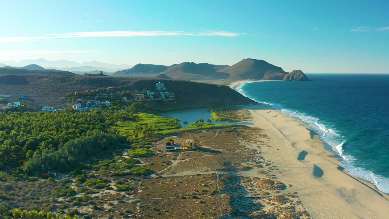 Aerial frame in Todos Santos with the beach running beside the ocean, a lagoon behind the sand, green vegetation at left, scattered buildings, and no visible people.