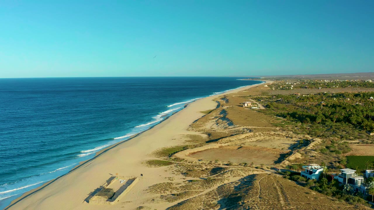 Aerial view of the Todos Santos shoreline with white wave bands closer to the sand, dunes crossing the center, small coastal buildings on the right, and no visible people.