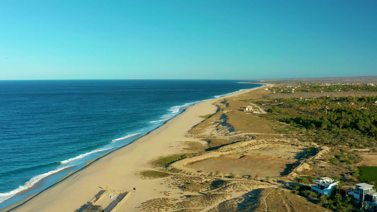 Opening aerial view over Todos Santos, Baja California Sur, with blue ocean at left, a long sandy beach, dune tracks, inland scrub, scattered houses, and no visible people.