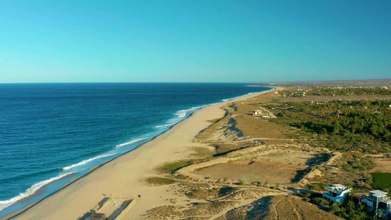 Aerial frame in Todos Santos showing the shoreline stretching into the distance, surf breaking along the beach, scrub-covered dunes inland, a few houses at the lower right, and no visible people.