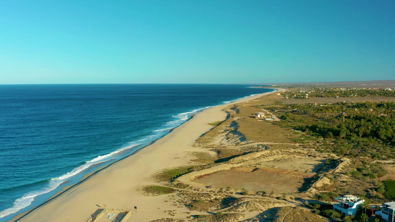 Wide aerial beach view with waves, dunes, scrubland, and houses inland.