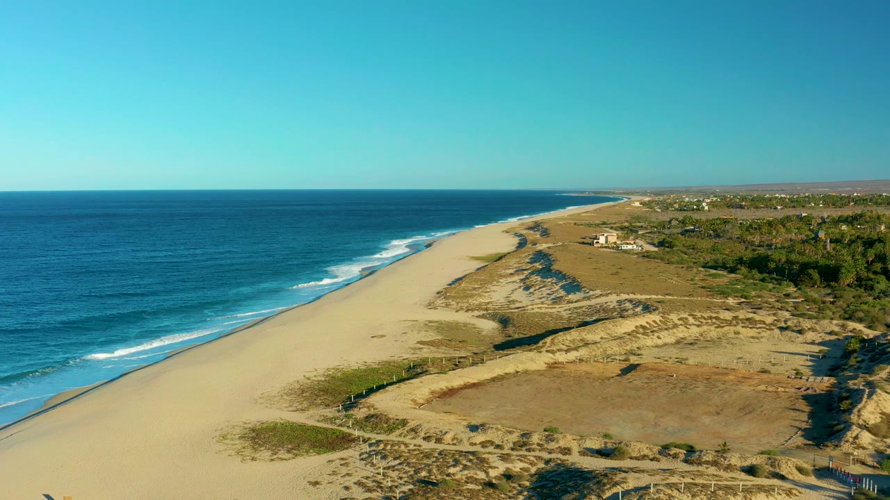 Early frame over Todos Santos coastline showing the beach running into the distance, small wave breaks along the left edge, dry dune mounds and scrub in the midground, and no visible people.