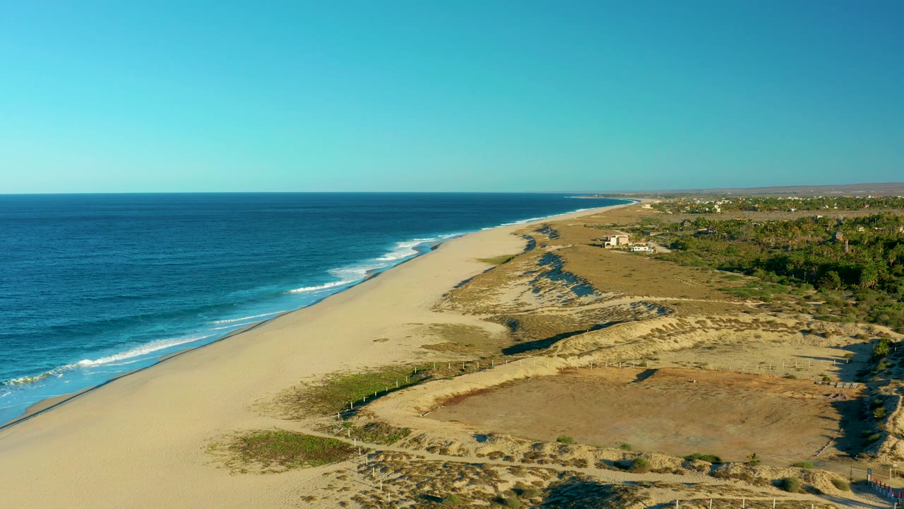 Aerial frame from Todos Santos with the shoreline angled from lower left toward the far distance, white surf tracing the beach, dune ridges and sparse grass inland, and no visible people.