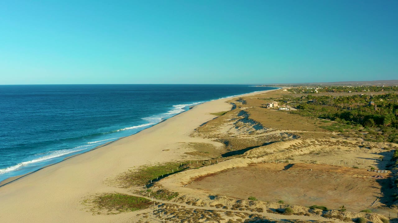 Mid-clip aerial view in Todos Santos with calm ocean at left, a broad empty beach, dry dune fields and scrub in the foreground and midground, distant coastal buildings, and no visible people.