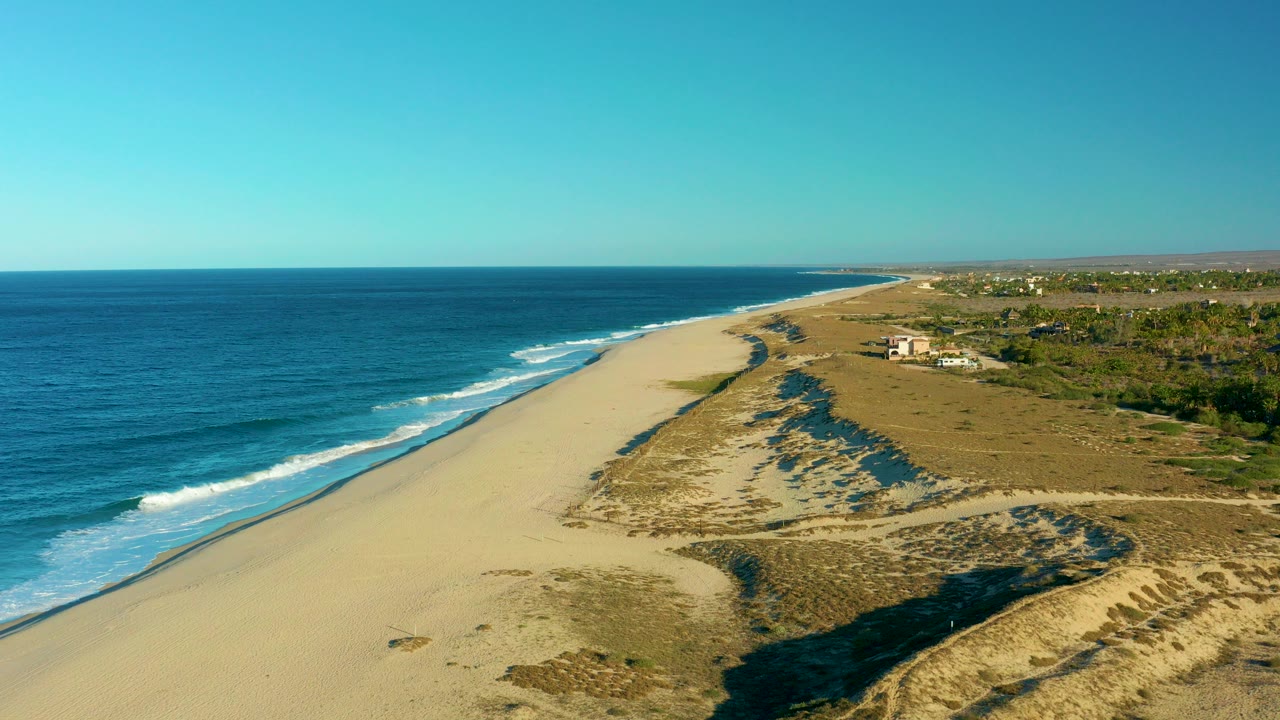 Aerial view over Todos Santos, Baja California Sur, with blue ocean on the left, a wide sandy beach, scrub dunes, inland vegetation, and no visible people.