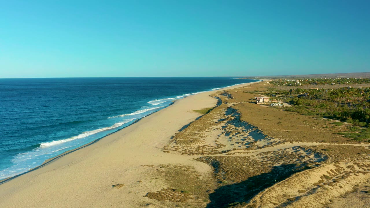 The Todos Santos shoreline stretches into the distance with white wave lines approaching the beach, dune shadows across the sand, inland greenery, and no visible people.