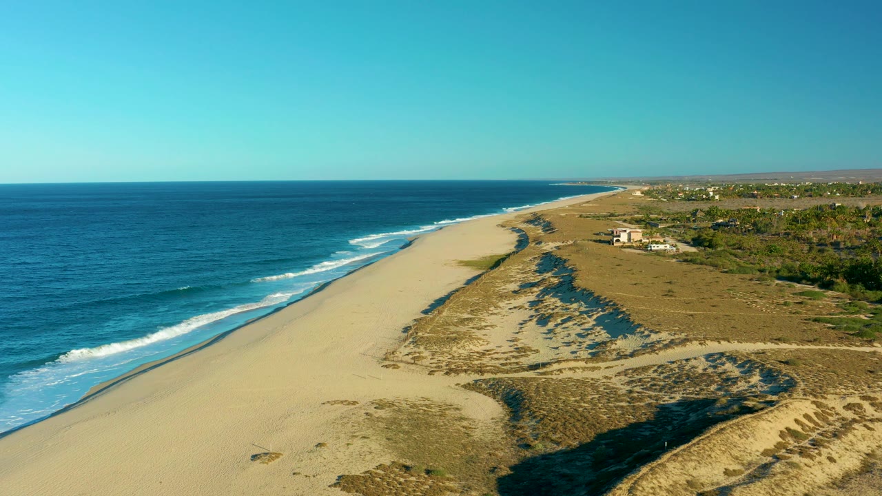 A high oblique aerial frame shows Todos Santos beach with the ocean left of frame, pale sand in the center, scrub-covered dunes to the right, distant buildings, and no visible people.