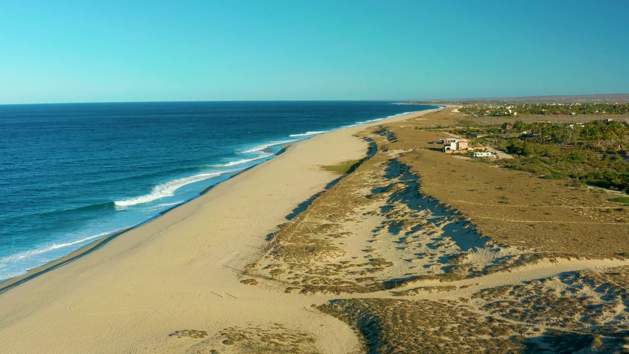 Aerial view over Todos Santos, Baja California Sur, showing blue ocean, white surf, a broad sandy beach, dune vegetation, distant structures, and no visible people.