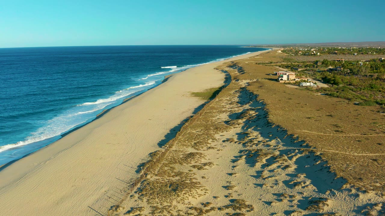 Aerial view in Todos Santos, Baja California Sur, with the shoreline running into the distance, waves breaking along the left edge, and dune vegetation casting shadows, with no visible people.