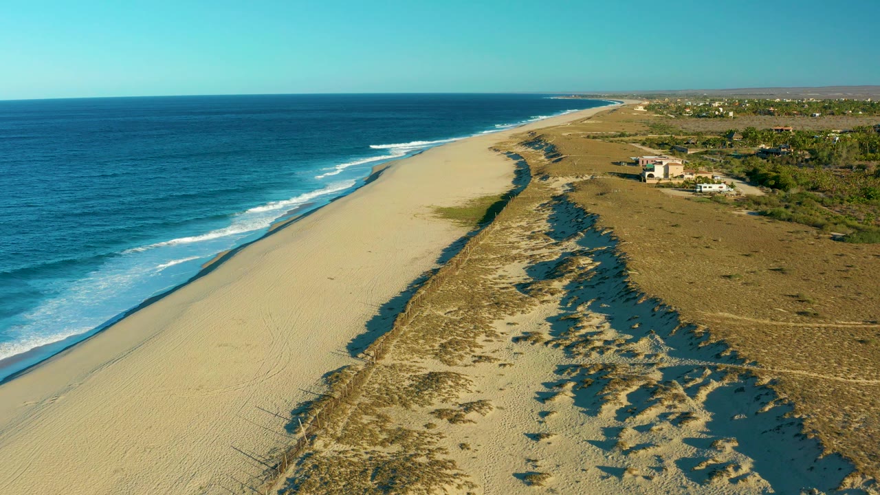 High coastal view of waves, open sand, dune shadows, and scrubland.