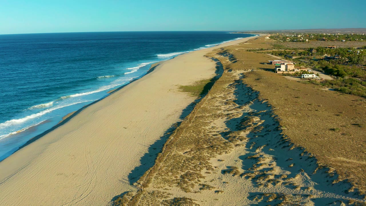 Aerial frame in Todos Santos showing white wave lines reaching the beach, a broad sand strip, scrub-covered dunes on the right, distant coastline, and no visible people.