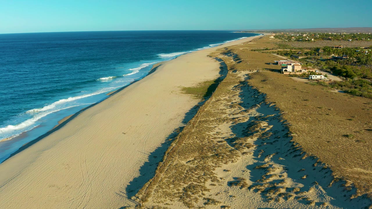 Drone-height view over Todos Santos with deep blue water, a narrow band of white surf, wide pale beach, darker dune shadows, scattered vegetation, inland buildings, and no visible people.