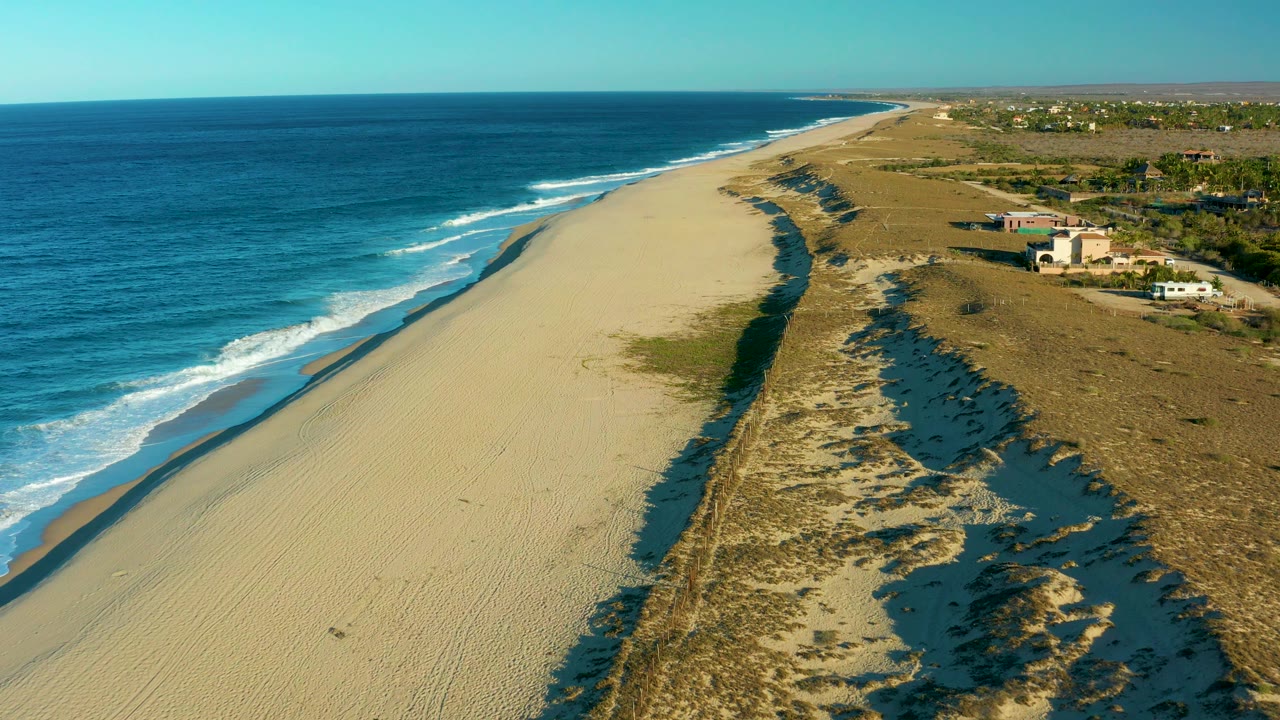 Drone-height view along the Todos Santos beach, with white waves rolling diagonally onto the sand, darker dune cuts holding water, inland scrub and small buildings in the distance, and no visible people.