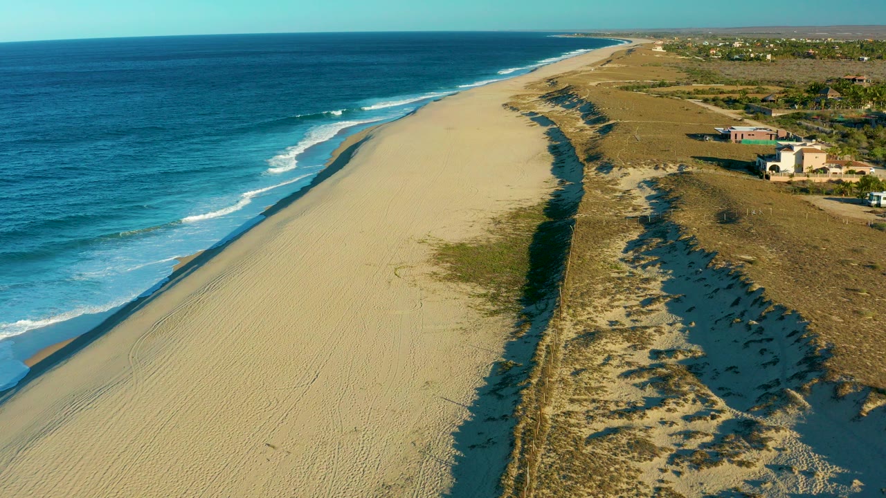 Aerial frame over Todos Santos beach with blue ocean on the left, a broad sandy shore, dune scrub on the right, distant coastal buildings, and no visible people.