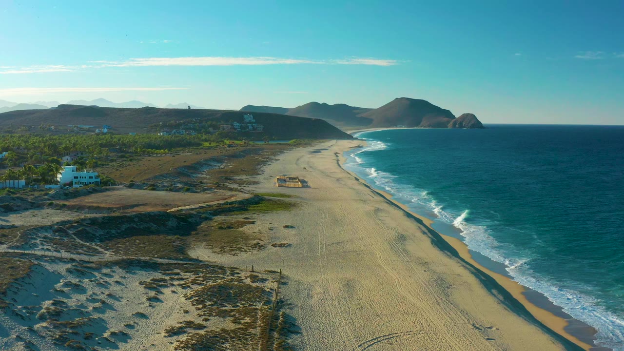 Wide aerial view over Todos Santos, Baja California Sur, with sandy beach in the center, blue ocean and surf on the right, dunes and a white building on the left, distant mountains, and no visible people.