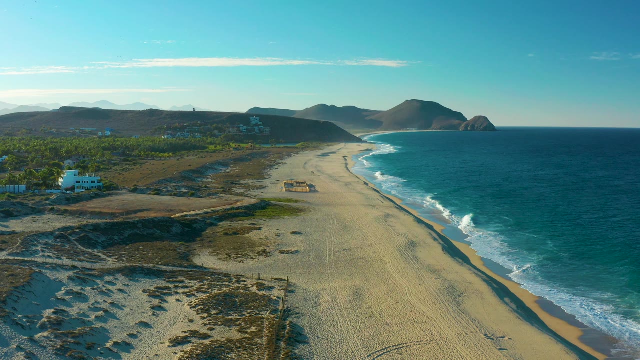 The Todos Santos coast appears from above with pale sand running diagonally, deep blue water on the right, a white building and green trees to the left, distant mountain shapes, and no visible people.