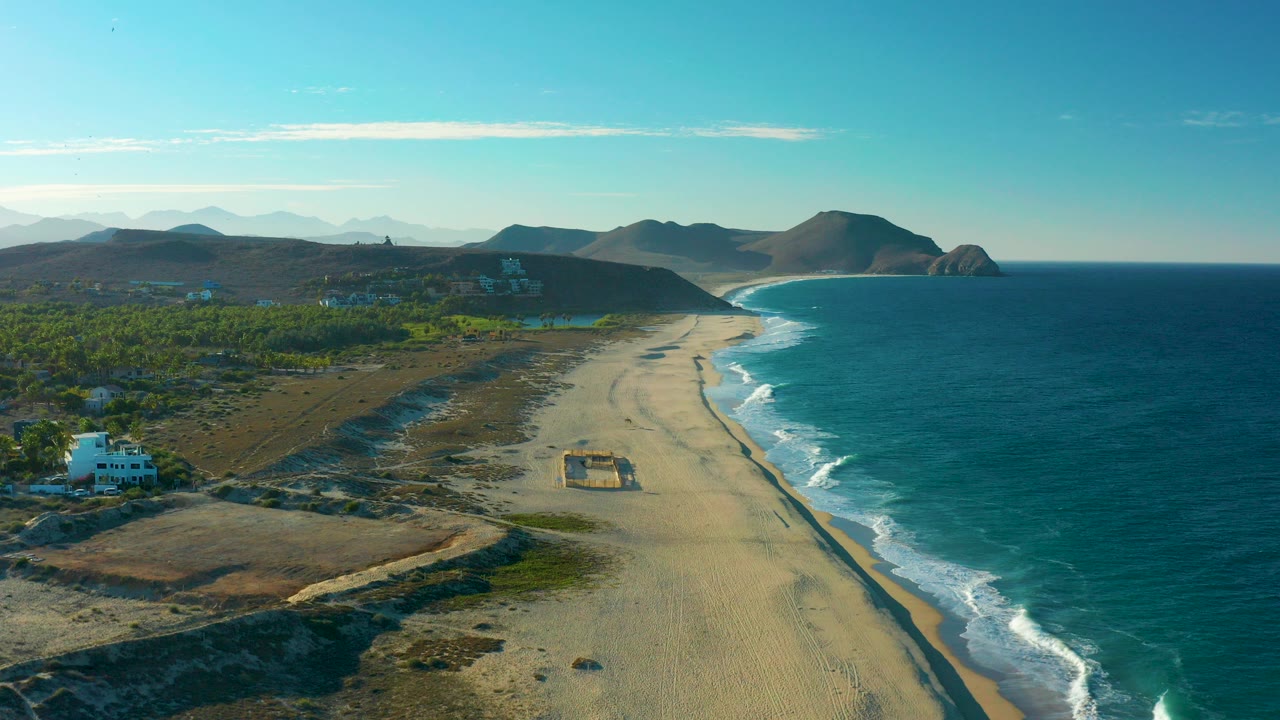 Opening aerial frame over Todos Santos, Mexico, with a sandy beach running toward distant mountains, blue ocean on the right, inland greenery on the left, a white building near the lower left, and no visible people.