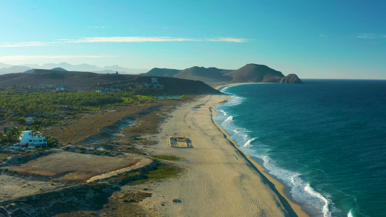 Second aerial frame of Todos Santos coastline, with the beach and wave line extending diagonally, the white building slightly lower at left, dry inland terrain, distant mountains, and no visible people.