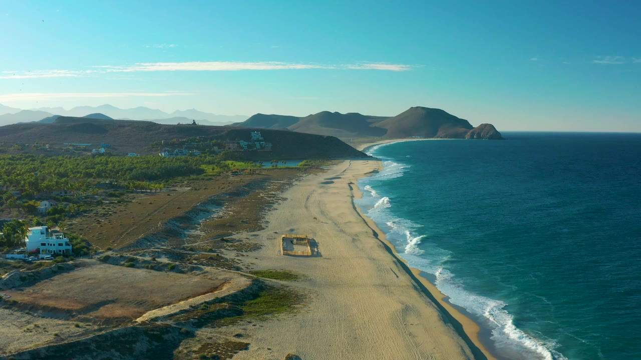 Third frame over the Todos Santos beach, showing more inland greenery and dry ground at left, empty sand through the center, blue ocean and surf on the right, distant mountains, and no visible people.