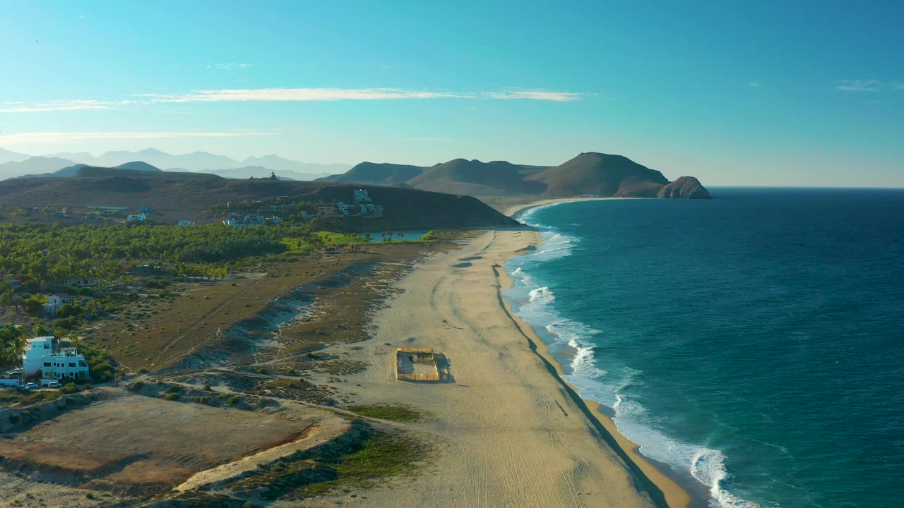 Coastal aerial view with white building at lower left, sandy beach center, ocean right, and mountains in distance.