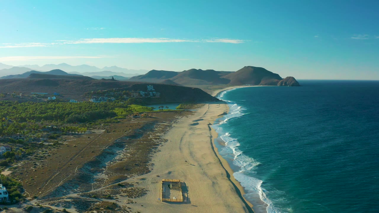 Aerial view in Todos Santos showing the sandy shore running toward a headland, white waves along the waterline, green vegetation inland, hillside structures, distant mountains, and no visible people.