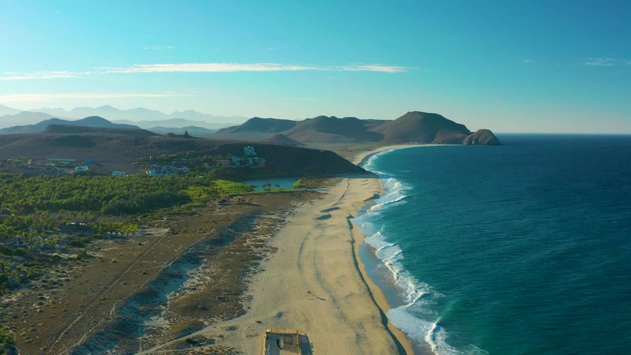 Mid-clip aerial frame over Todos Santos with a long tan beach below, waves breaking on the right edge, inland greenery and hillside buildings on the left, hazy mountain layers beyond, and no visible people.