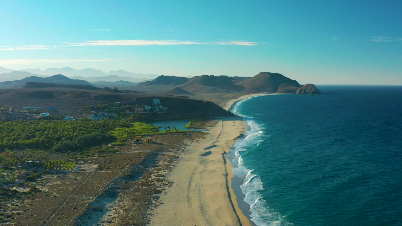 Wide aerial view over Todos Santos, Baja California Sur, with a long sandy beach between blue ocean and lagoon water, green vegetation inland, distant hills, clear sky, and no visible people.