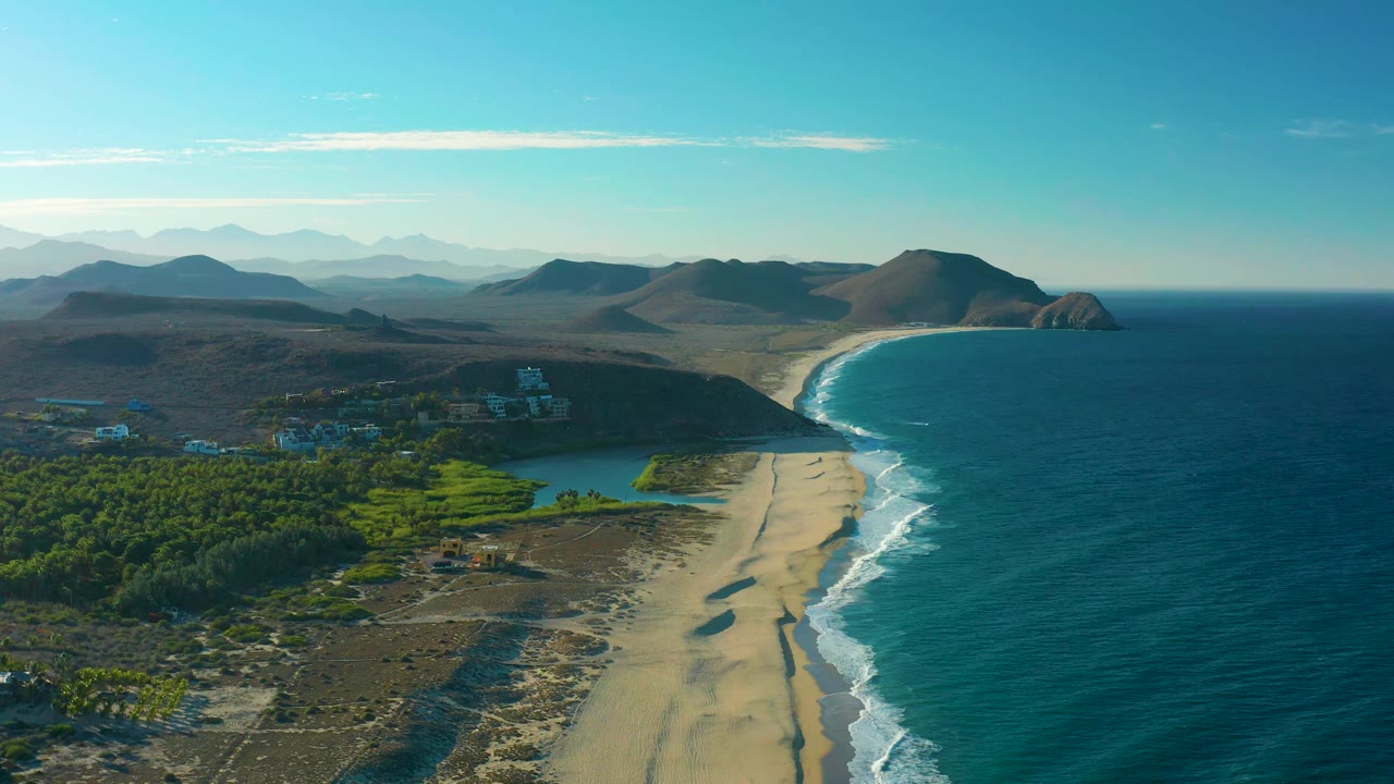 Wide aerial frame in Todos Santos showing the bright sand strip, white waves along the right edge, lagoon water beside green vegetation, rounded hills, distant ridges, and no visible people.