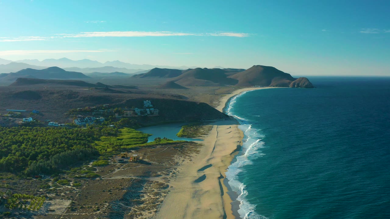 Elevated Todos Santos coastline view with dense green palms in the foreground, a turquoise lagoon, sandy beach, white wave bands, a rounded headland, hazy mountains, and no visible people.