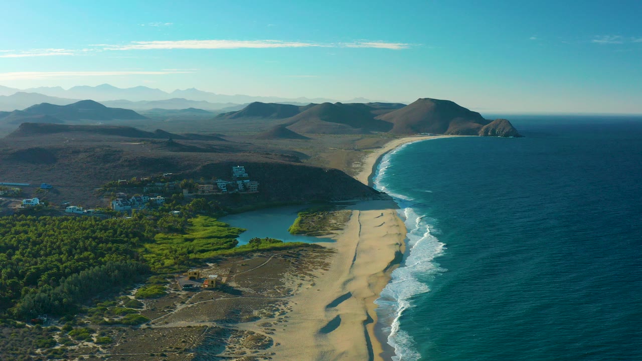 Opening aerial frame over the Todos Santos coast with a sandy beach, white surf, blue ocean, green lagoon area, dry hills, distant mountains, and no visible people.