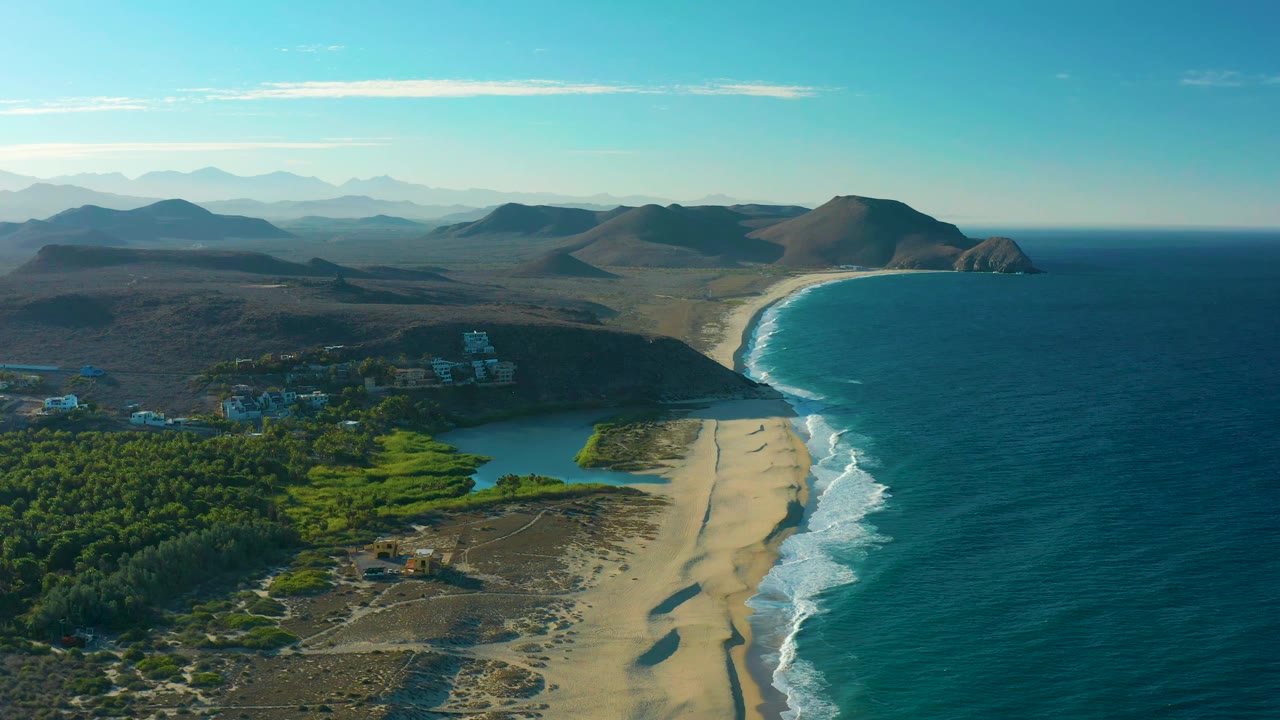 Wide aerial coastline with waves, sand, lagoon, greenery, and distant hills.