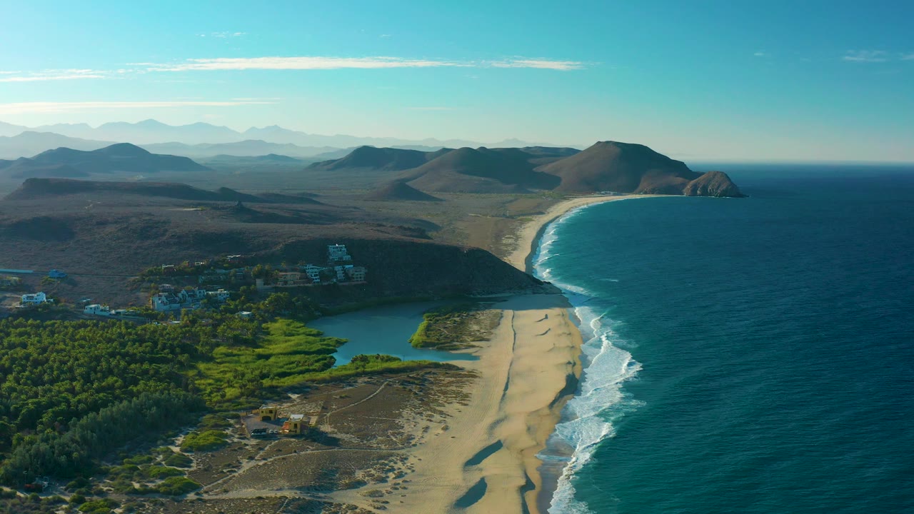 High coastal view with beach, surf, lagoon, green trees, and mountain backdrop.