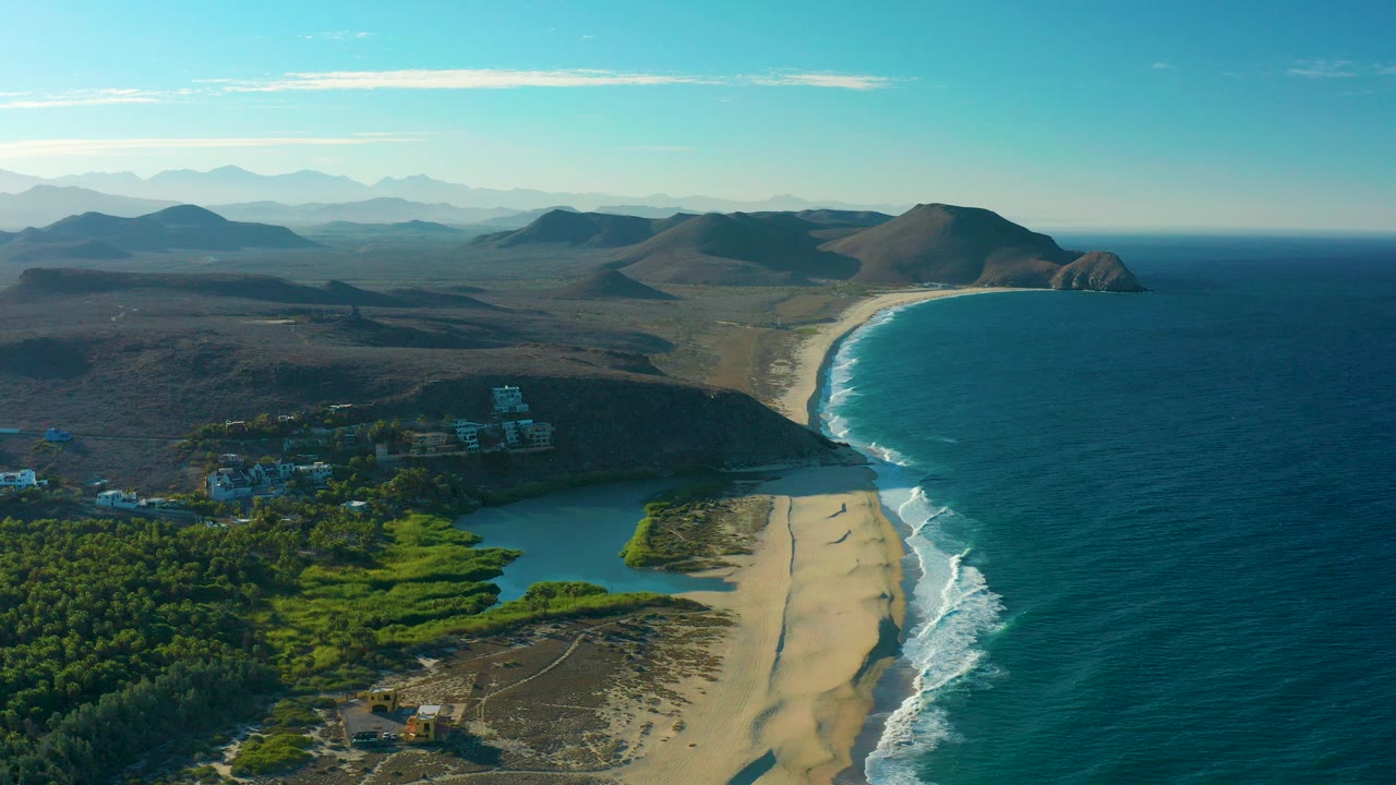 Opening aerial frame over Todos Santos, Baja California Sur, Mexico, showing a green lagoon beside a sandy beach, white ocean surf, low hills, distant mountains, clear blue sky, buildings near the water, and no visible people.