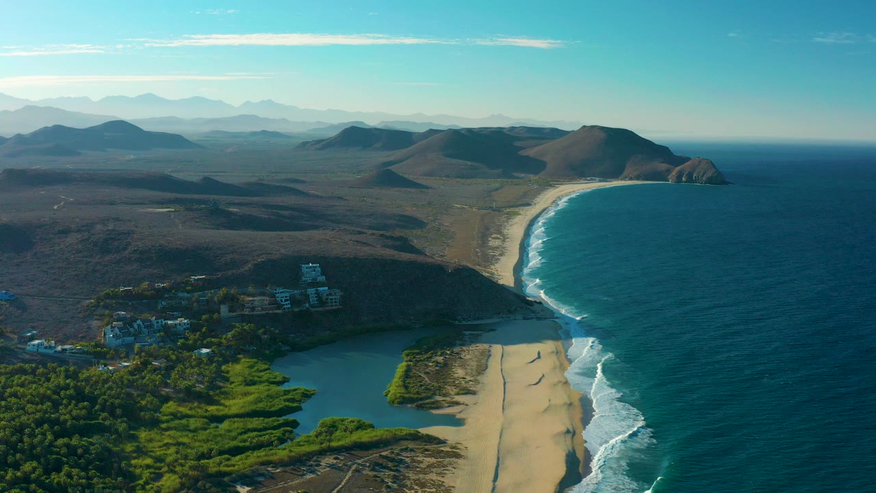 Aerial view over Todos Santos, Baja California Sur, Mexico, with green lagoon vegetation in the foreground, a sandy beach curving beside blue ocean, distant hills, and no visible people.