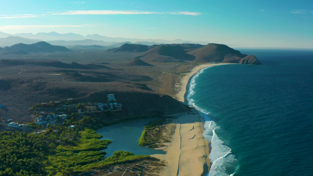 Wide aerial frame in Todos Santos with the surf line running along a broad sandy beach, lagoon water and greenery below, dry hills beyond the shore, and no visible people.