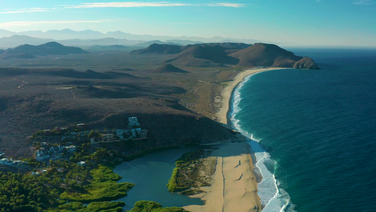 Aerial coastline with lagoon, sandy beach, ocean waves, buildings, and distant hills.